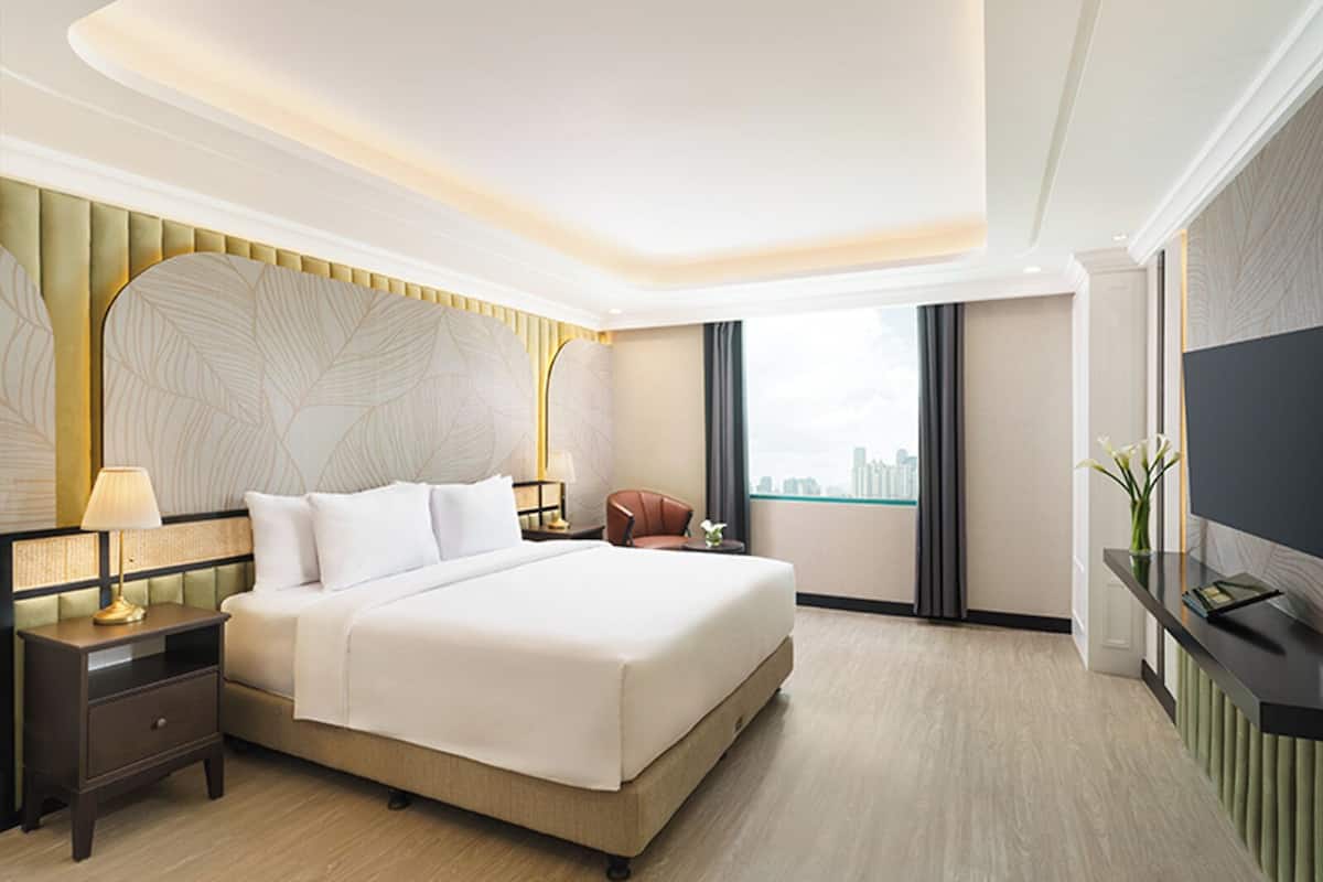 A modern hotel room with a large bed, a nightstand with a lamp, a seating area with a chair, a television on a stand, and a view of the city through the window.