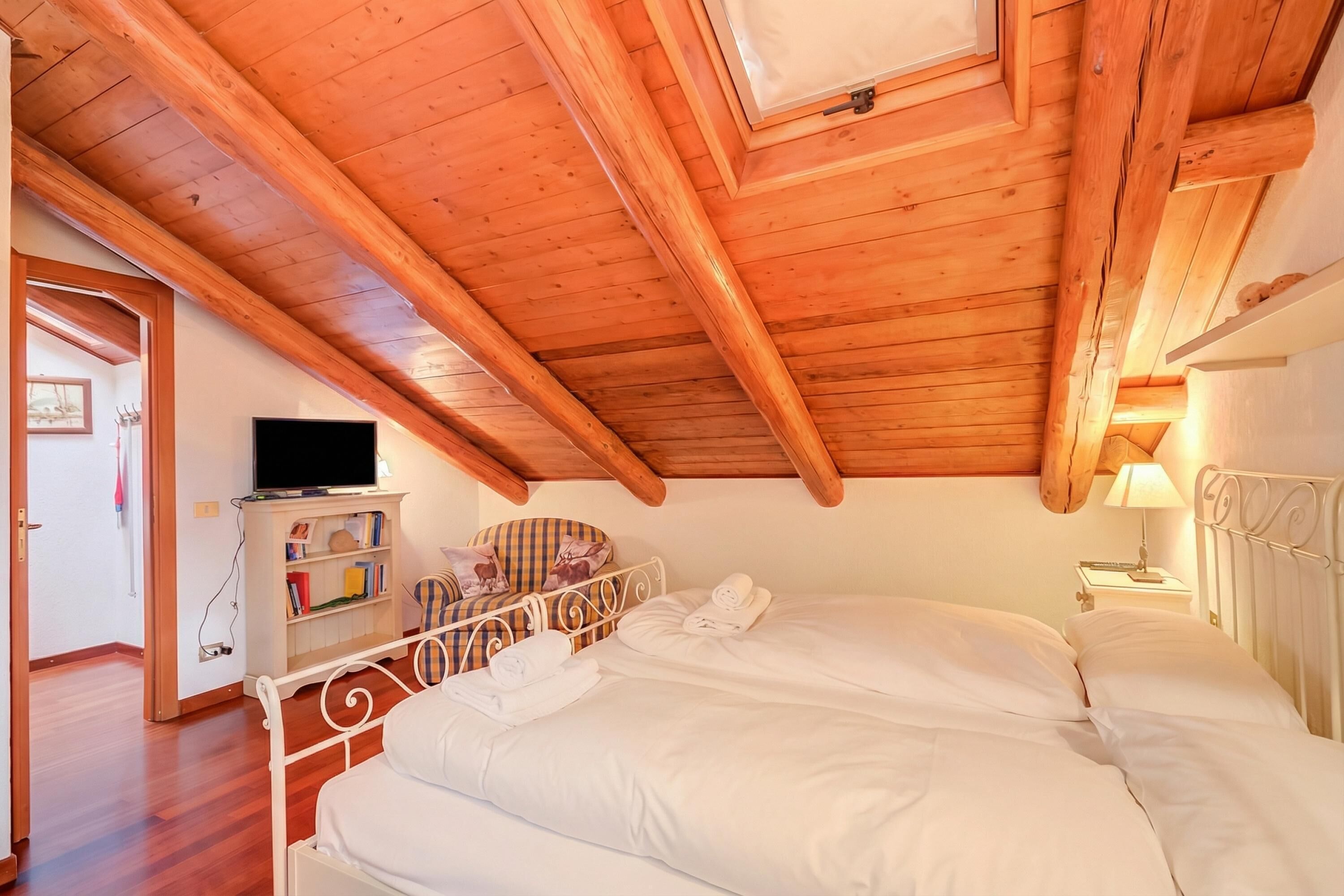 Une chambre avec un plafond en pente, un lit recouvert de draps blancs, une table de chevet avec une lampe et un téléviseur posé sur un meuble.