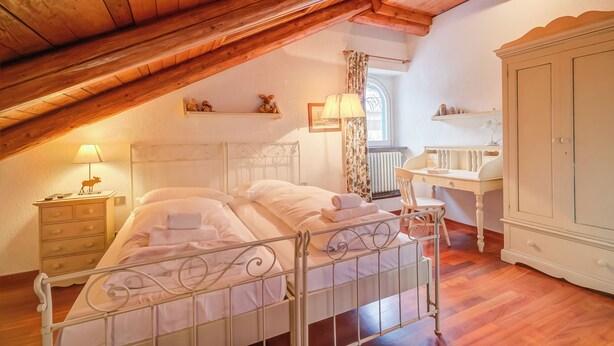 A bedroom with a sloped ceiling, a bed with white linens, a nightstand with a lamp, a dresser, and a window with curtains.