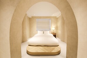 Un dormitorio con una cama centrada debajo de un amplio ventanal arqueado, una mesita de noche con una lámpara, y una plataforma de madera al pie de la cama.