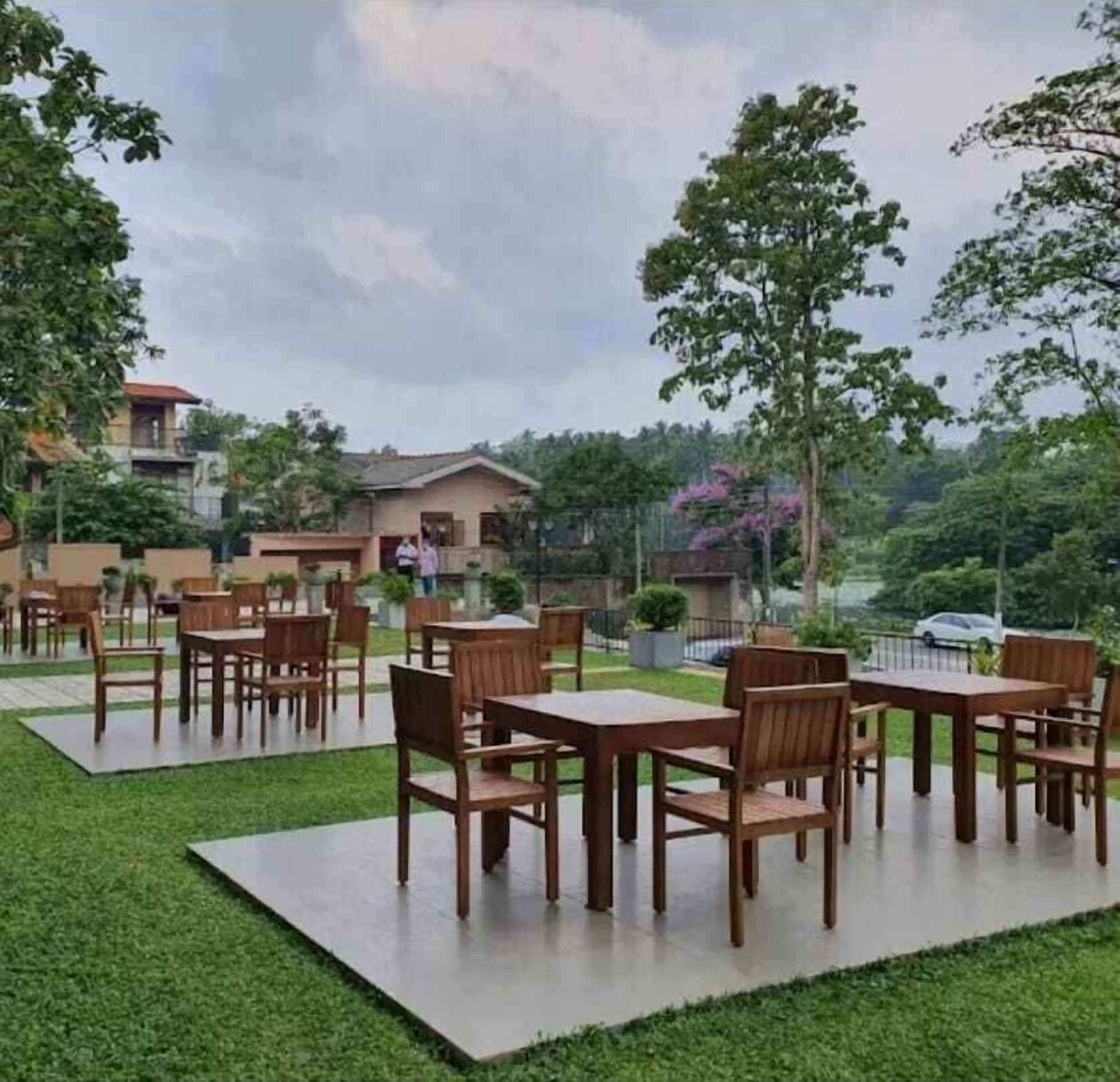 Un patio exterior con varias mesas y sillas de madera, rodeado de césped cuidado y árboles.