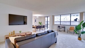 Un salon moderne avec un canapé gris, un téléviseur à écran plat fixé au mur, un coin repas comprenant une table et des chaises, et de grandes fenêtres offrant une vue sur la ville.