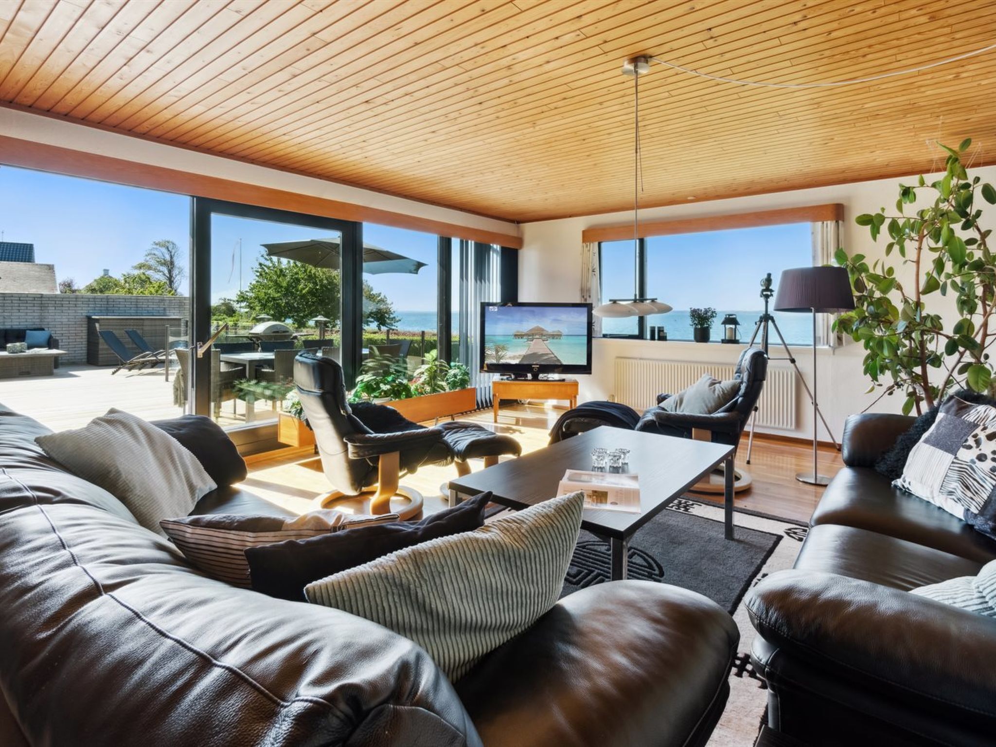 A modern living room with leather sofas, a coffee table, and a TV. There is a large window offering a view of the outdoors.