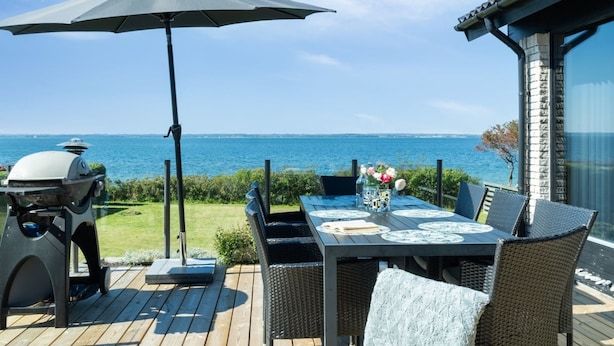 An outdoor dining area with a view of the sea, a grill, and a table set with plates and a vase of flowers.