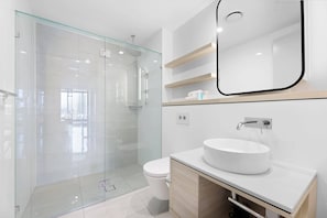 A modern bathroom with a glass shower enclosure, a white sink, and a mirror with a wooden shelf above. - Luxe Oceanfront 1-Bed High Rise with Pool & Gym (Surfers Paradise)