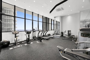 A modern gym with cardio machines, weightlifting equipment, and large windows offering a view of the city. - Luxe Oceanfront 1-Bed High Rise with Pool & Gym (Surfers Paradise)