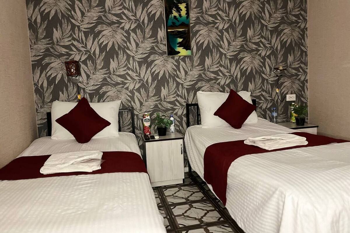 A hotel room with two beds, each with a red pillow and white bedding. There is a small table with a bottle and a plant, and a painting on the wall.