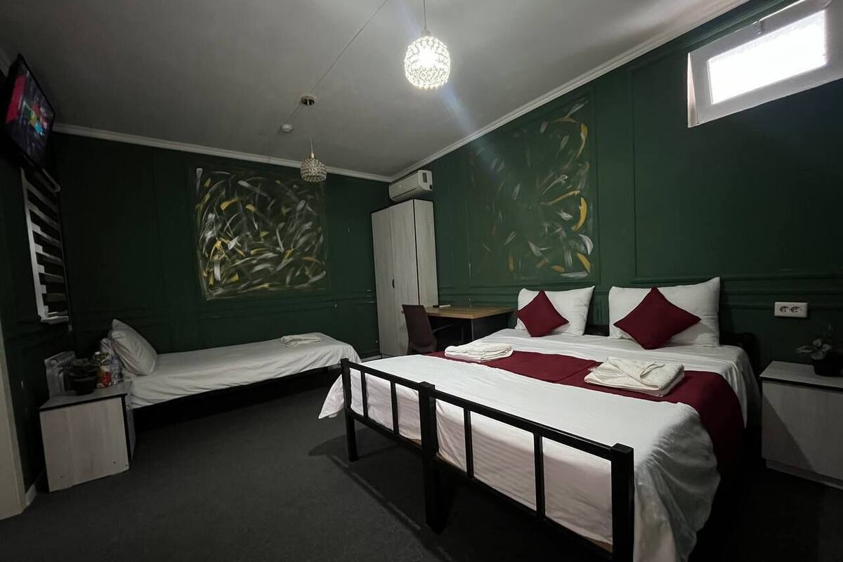 A room with two single beds, a desk, and a chair. There is a window on the right wall and an air conditioning unit on the left wall.