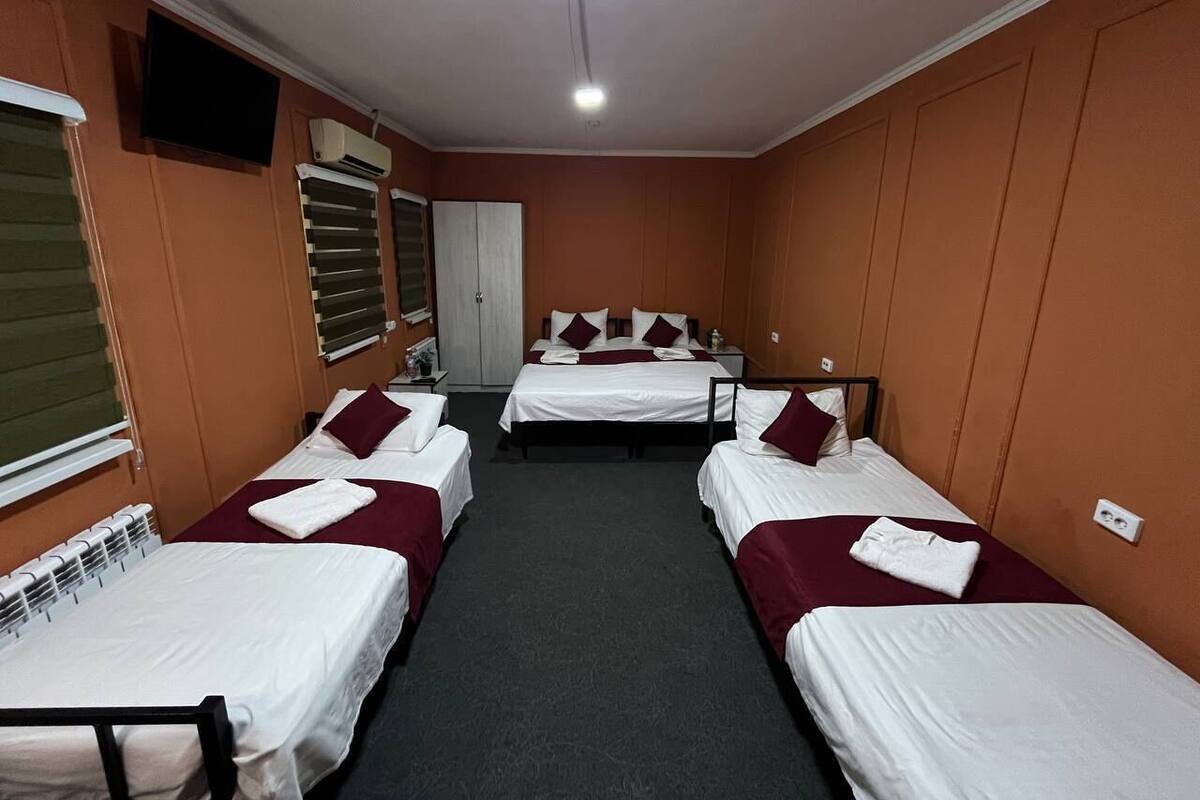 A hotel room with three single beds, each with white linens and maroon pillows. There is a television on the wall, a window with blinds, and a door.