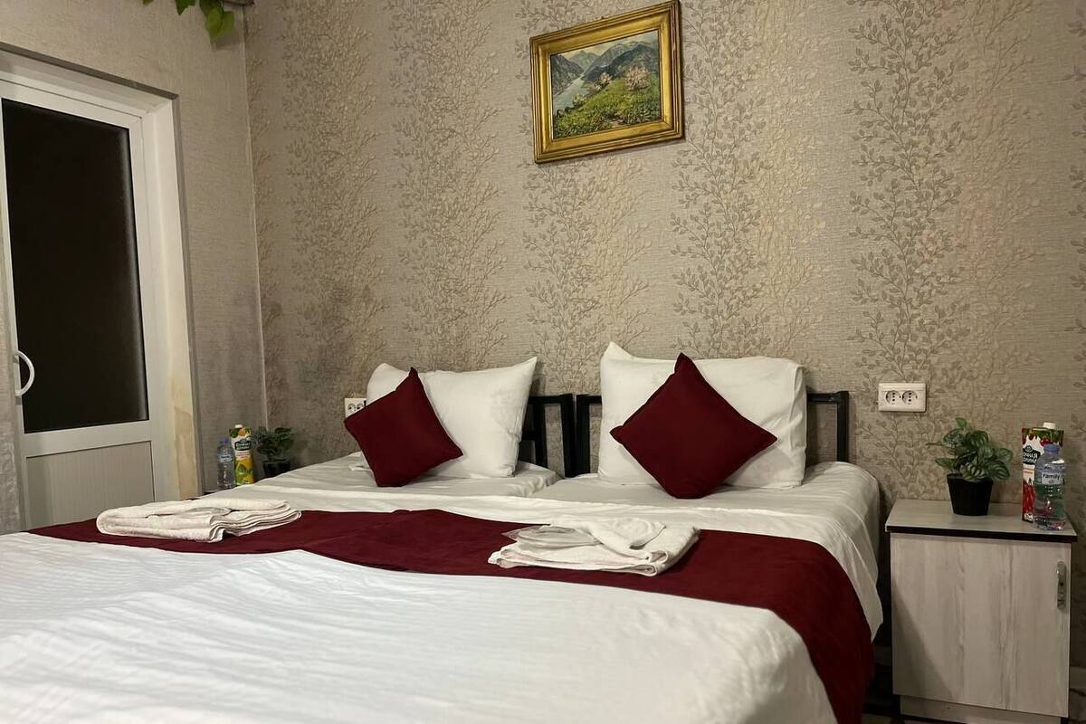 A hotel room with a bed, two pillows, a nightstand, a plant, and a painting on the wall.