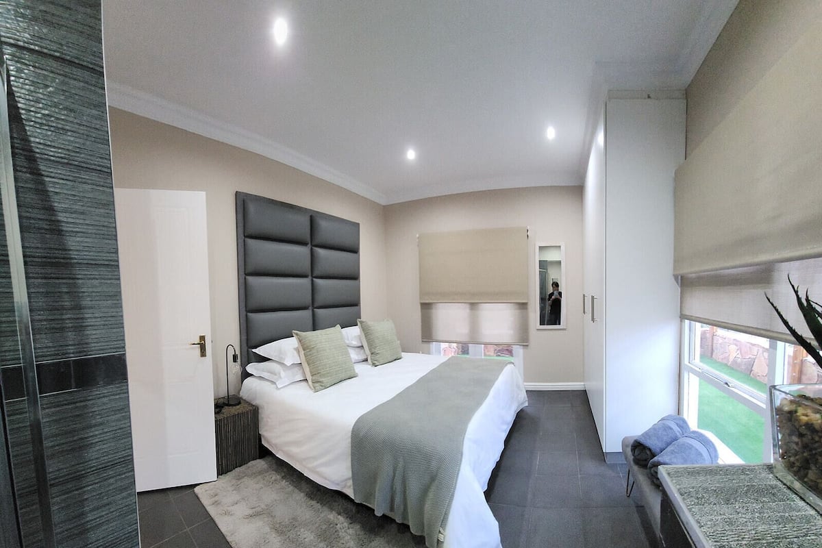 A modern bedroom with a large bed, a bedside table, a nightstand, a mirror, and a window with a view.