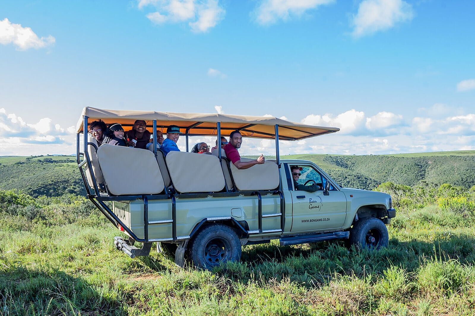 Vehículo safari con techo, transportando pasajeros, estacionado en un campo de césped con un paisaje colinas al fondo.