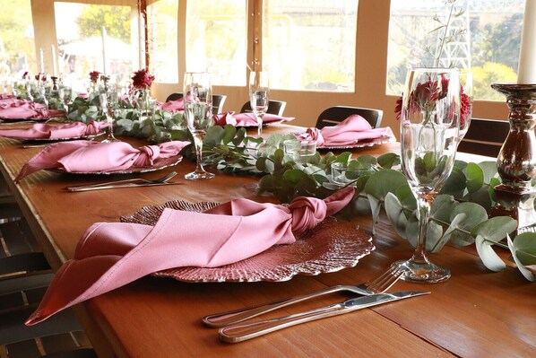 A long dining table set with pink napkins, clear wine glasses, and silverware, surrounded by greenery and a view of the outdoors. - Bon Game Reserve (Albertinia)