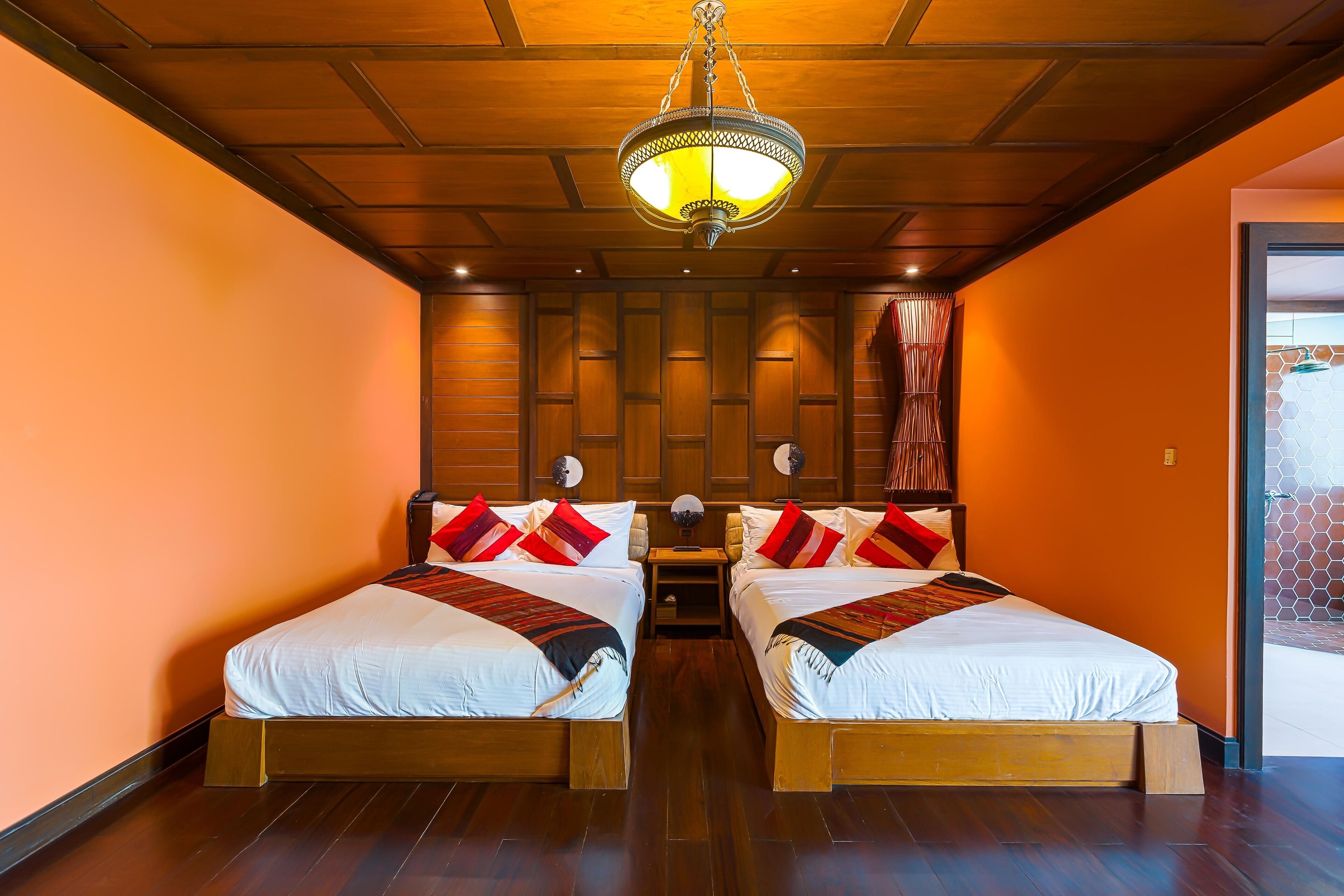 A double room with two beds, wooden headboards, a central chandelier, and a view of a brick wall.