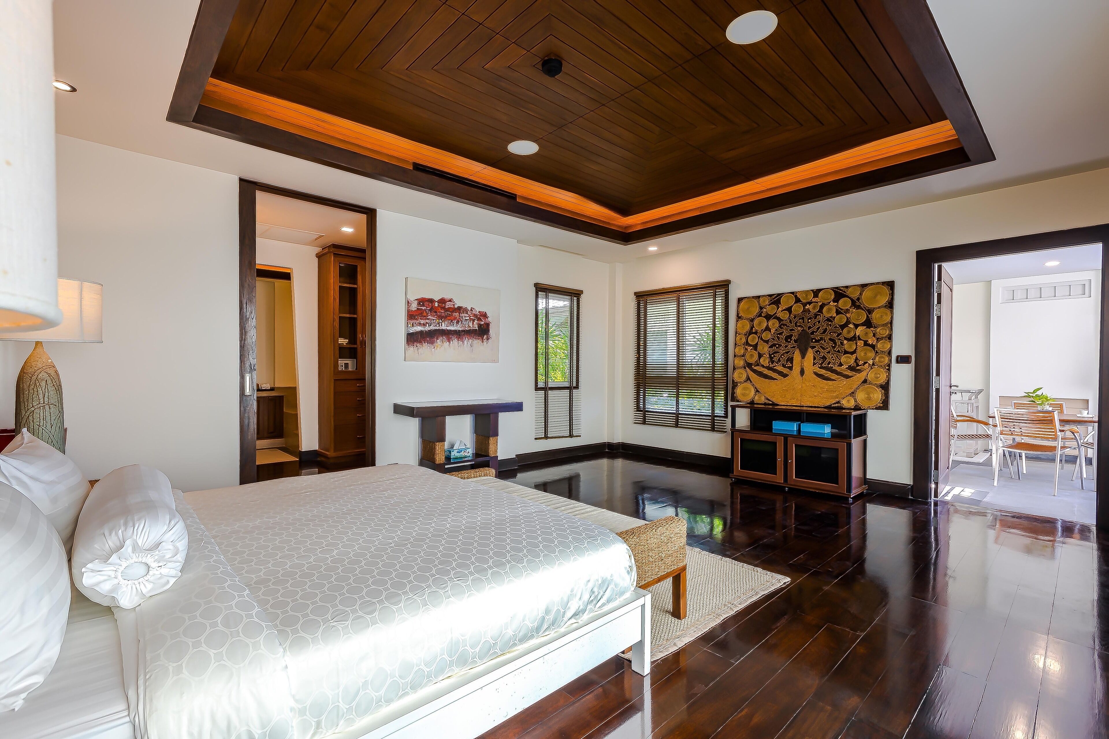 A modern bedroom with a large bed, wooden ceiling, and a dining area visible through an open door.