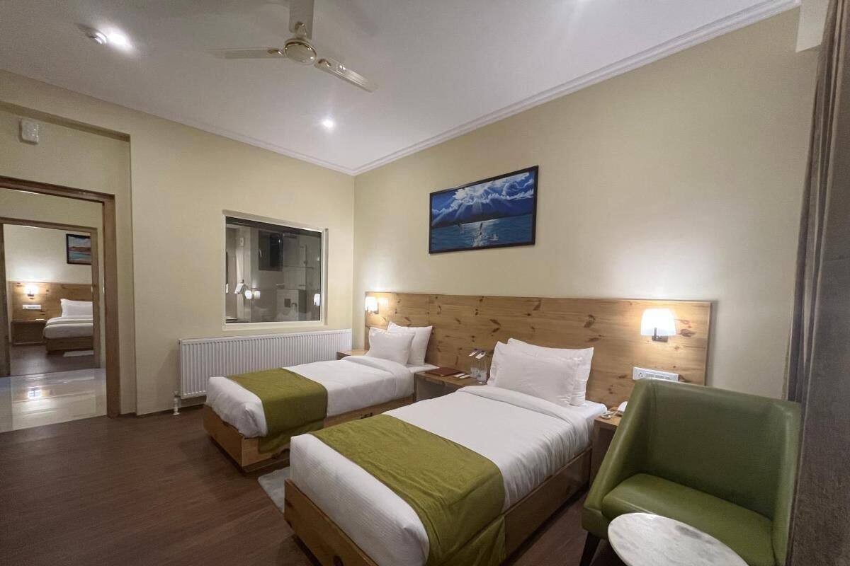 Habitación de hotel con dos camas, una silla verde, una mesita redonda, un espejo y un cuadro en la pared.
