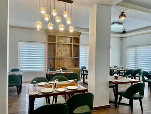 Zona pranzo con tavoli in legno e sedie verdi, una libreria in legno e lampade a sospensione.