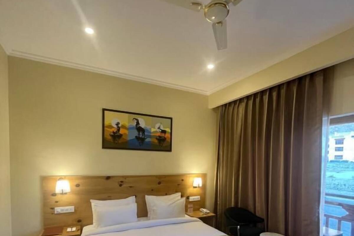 Habitación de hotel con una cama grande, ventilador de techo, un cuadro en la pared, una mesita de noche y una ventana con cortinas.