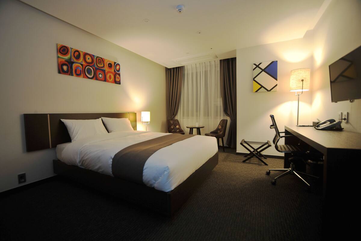 A hotel room with a large bed, a desk with a chair, a lamp, and a painting on the wall.