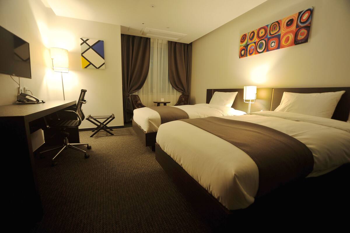 A hotel room with two beds, a desk with a chair, a TV, and a lamp.