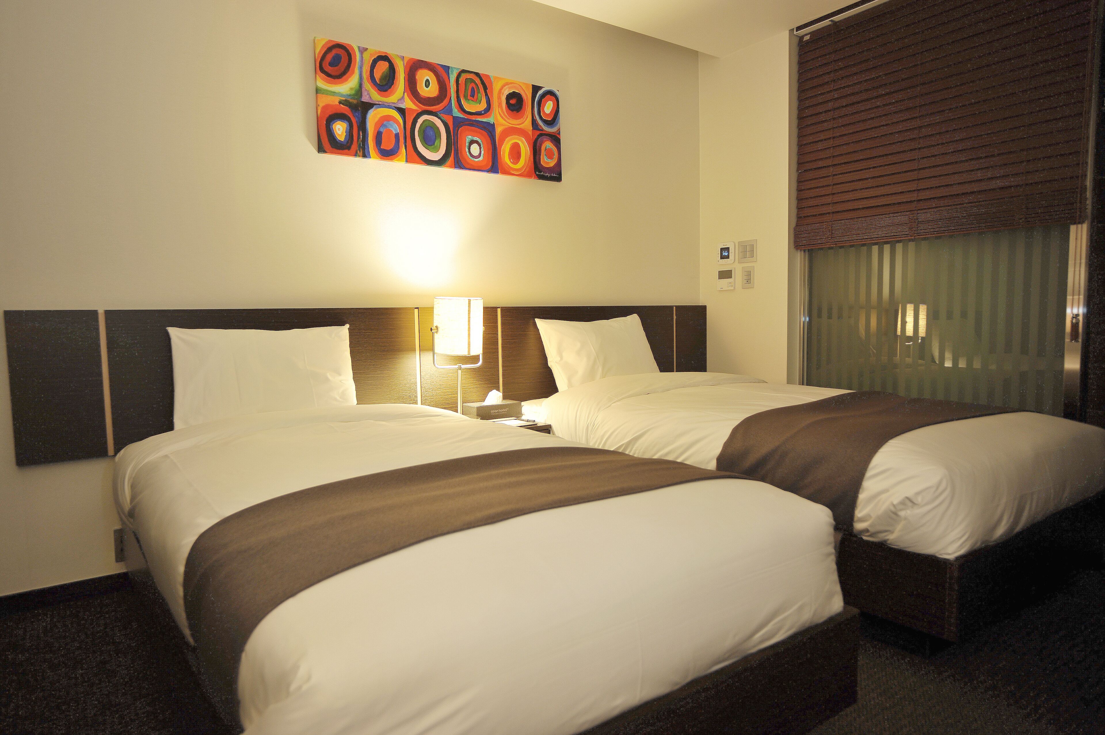 A hotel room with two beds, a headboard with a light fixture, a colorful abstract painting, and a window with blinds.