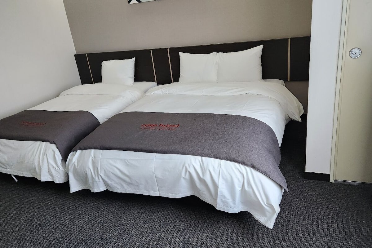 A double bed with white bedding and two pillows, a dark headboard, and a carpeted floor.