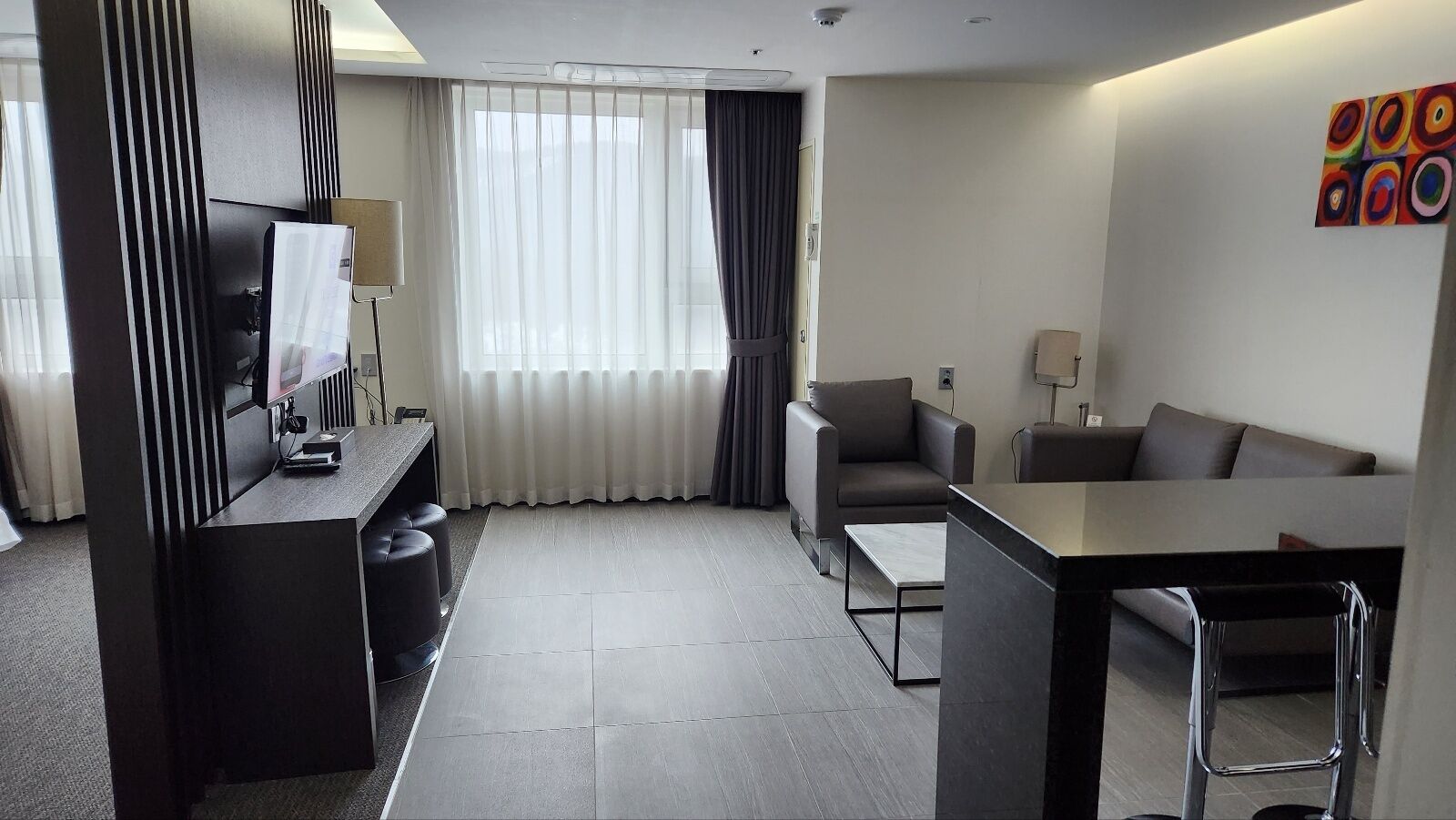 A modern hotel room with a desk, chair, sofa, and a large window with curtains.