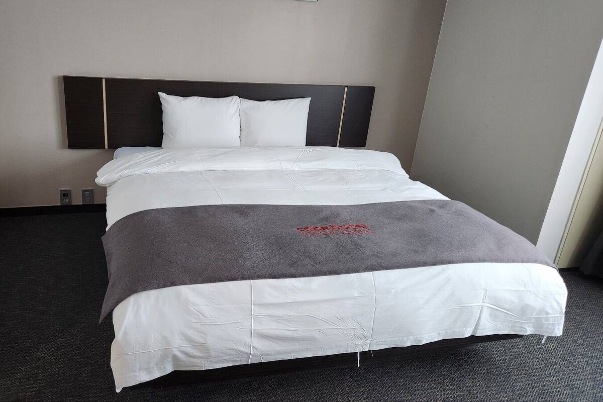 A hotel room with a large bed, dark headboard, and a grey blanket.
