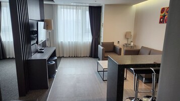 Una habitación de hotel moderna con comedor, sofá, televisor y un ventanal con cortinas.