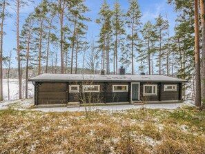 Lumihunnun peittämä hirsimökki, jonka ympärillä kohoaa korkeita mäntyjä.