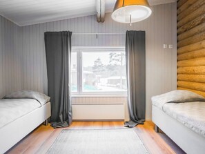 A room with two beds, a window with curtains, a wooden wall, and a ceiling lamp.