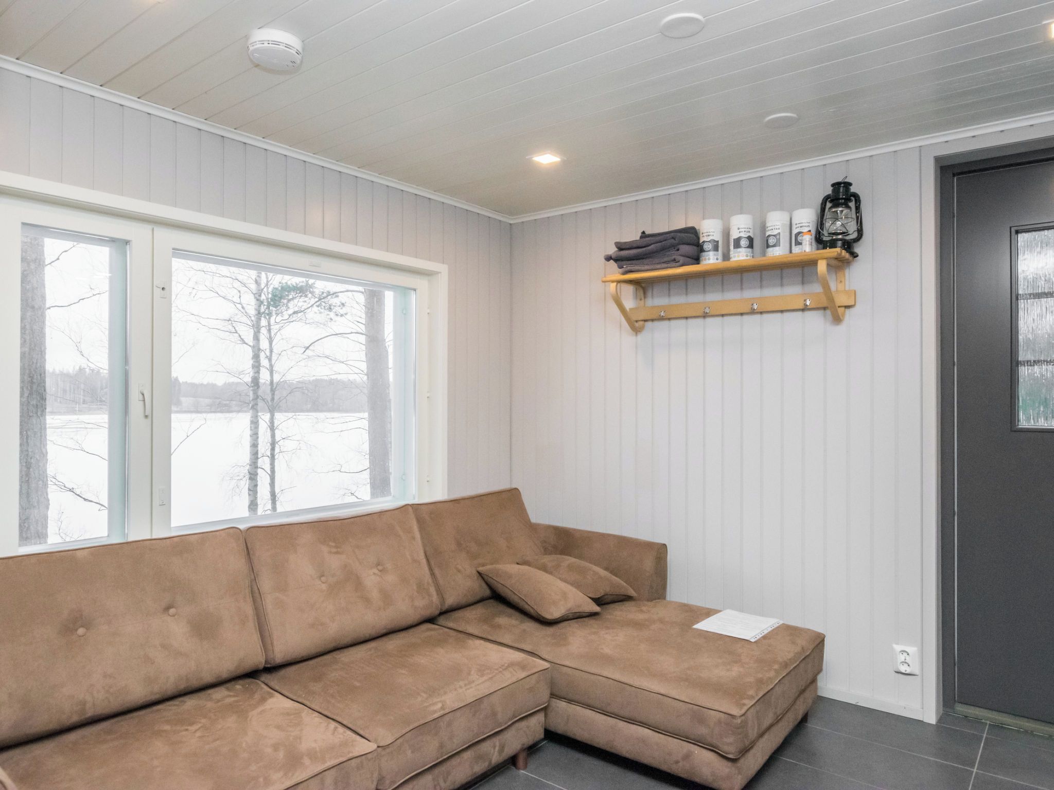 A room with a brown sectional sofa, a window with a view of trees, and a shelf with toiletries.