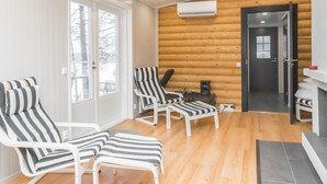 Een kamer met houten wanden, een witte deur en een open haard. Er staan gestreepte fauteuils en een klein tafeltje met een speaker.