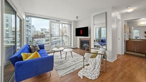 Een moderne woonkamer met een blauwe bank, een open haard, een televisie en uitzicht op de stad door grote ramen.
