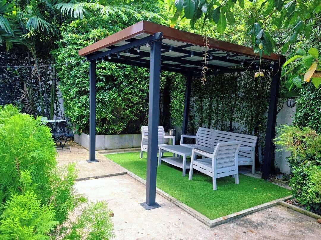 A covered outdoor seating area with a bench and table, surrounded by lush greenery.