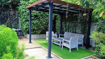 A covered outdoor seating area with a bench and table, surrounded by lush greenery.