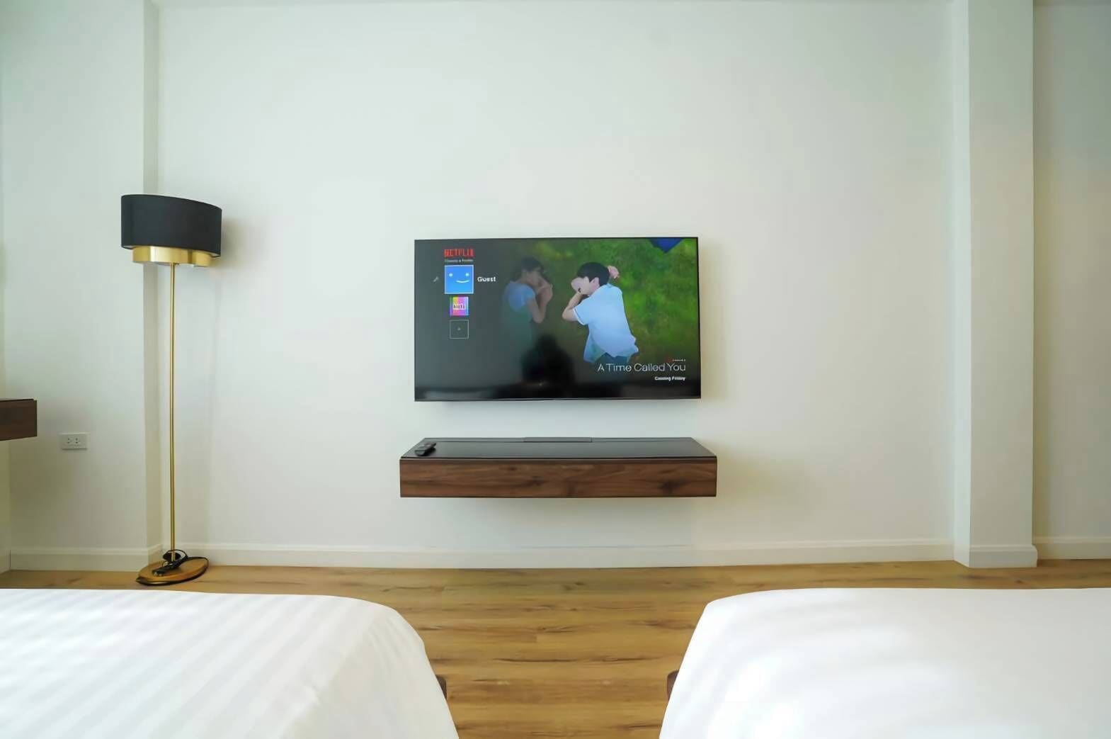 A hotel room with a flat-screen TV mounted on the wall, a bedside table, and a floor lamp.