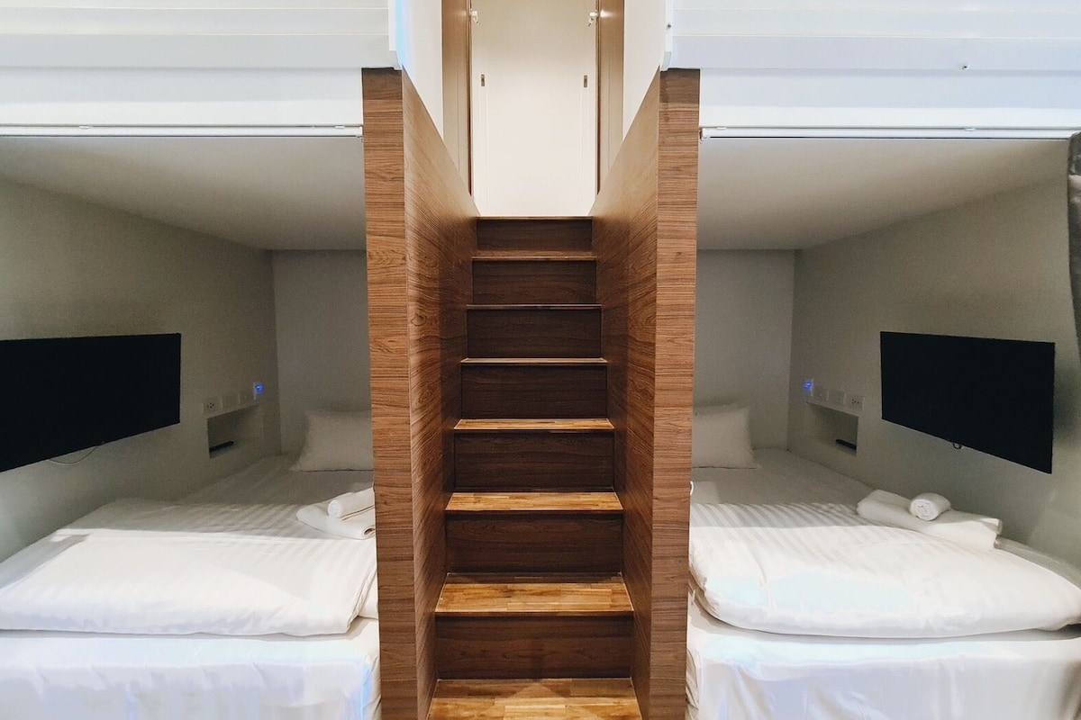 Habitación de hotel con una escalera central de madera que conecta dos camas, cada una con un televisor y un cojín.