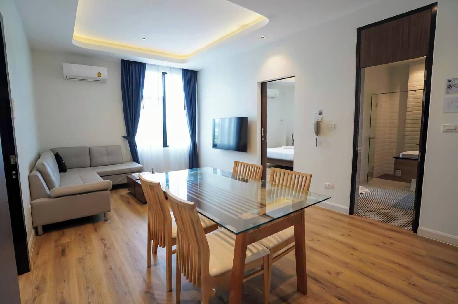 Un salon moderne avec une table à manger en verre, des chaises en bois, un canapé et un téléviseur fixé au mur.