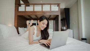 Una persona recostada en una cama con una computadora portátil, una estructura de literas al fondo y un cubo de basura a un lado.