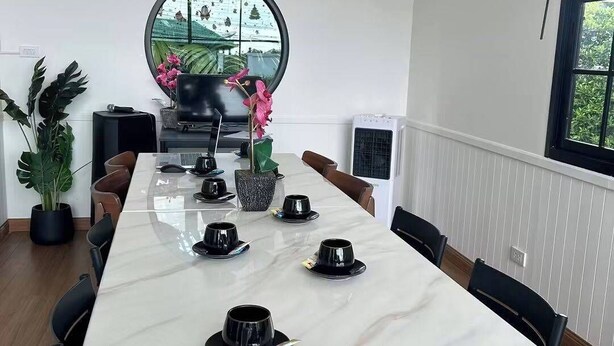 A modern dining area with a long table set for a meal, a large window with a view of greenery, and an air conditioning unit on the wall. - Play Poshtel & Cafe (Krabi)
