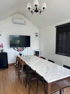 Un espace repas comprenant une longue table, des chaises, un téléviseur à écran plat, un appareil de climatisation et une fenêtre avec des stores.