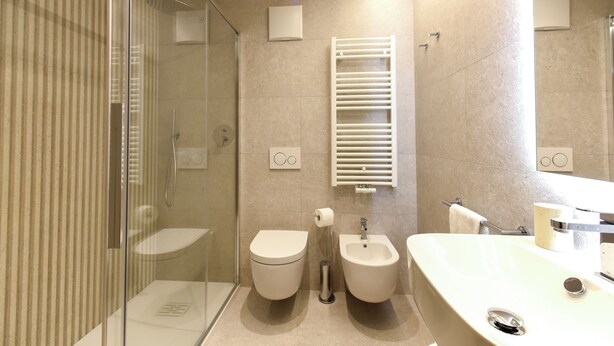 A modern bathroom with a glass shower enclosure, a toilet, a bidet, and a sink with a mirror. - Naturae Lodge (Alleghe)