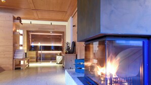A modern living room with a fireplace, wooden furniture, and a kitchen area in the background. - Naturae Lodge (Alleghe)