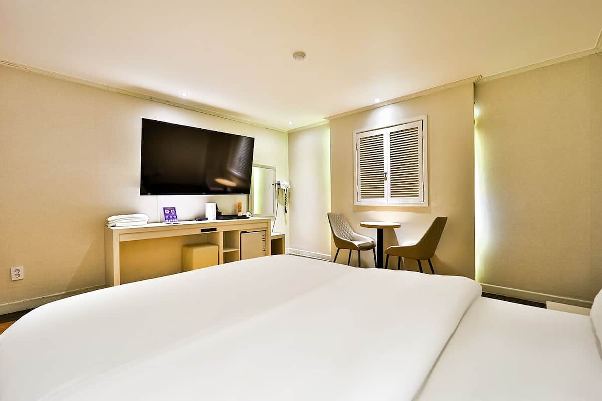 A hotel room with a large bed, a desk with a flat-screen TV, a chair, a small table, and a window with blinds.