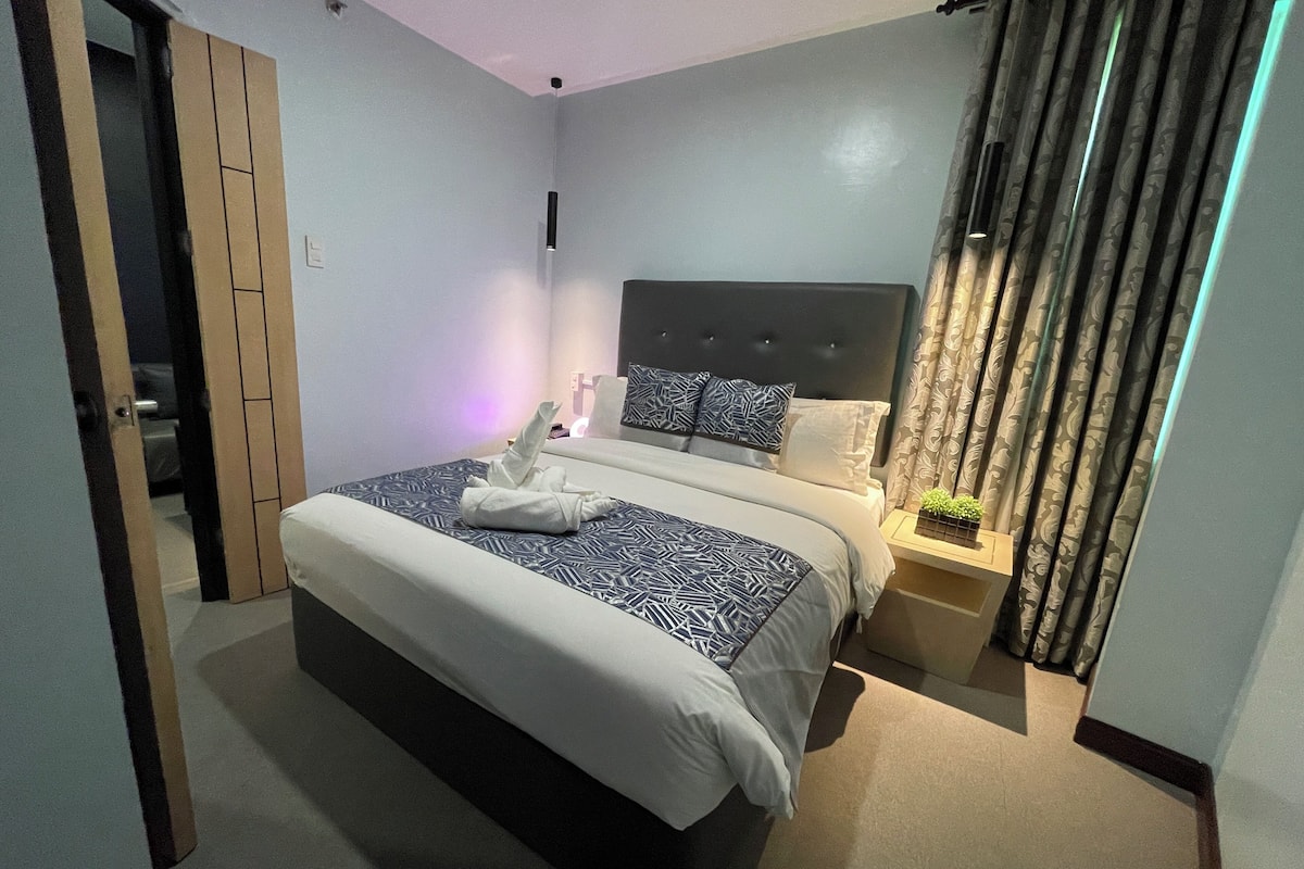 A modern hotel room with a large bed, a bedside table with a lamp, and a view of the outside through curtains.