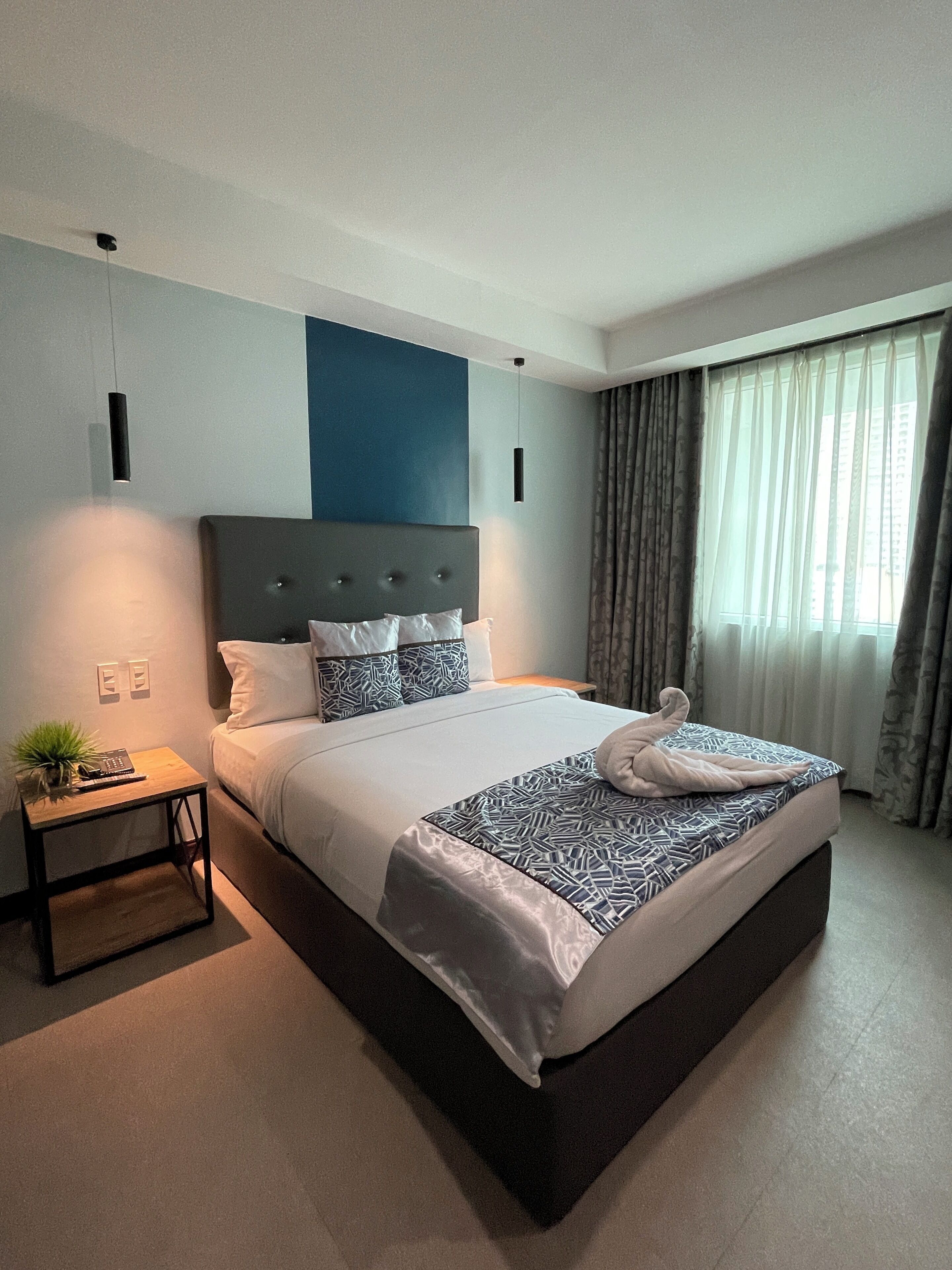 Habitación de hotel moderna con una cama grande, una mesita de noche con una planta y vistas a la ciudad a través de cortinas translúcidas.