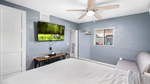 Une chambre à coucher comprenant un lit, un téléviseur fixé au mur, un ventilateur de plafond et une fenêtre avec des stores.