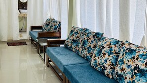 A modern living room with a blue sofa set, white curtains, and a visible bathroom with a sink and mirror. - Chaubey Farms- Relax & Rejoice
- The best farm stay in Hyderabad (Chilipched)