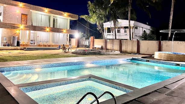 A swimming pool with a tiled edge, a building with a balcony and lights, and palm trees. - Chaubey Farms- Relax & Rejoice
- The best farm stay in Hyderabad (Chilipched)